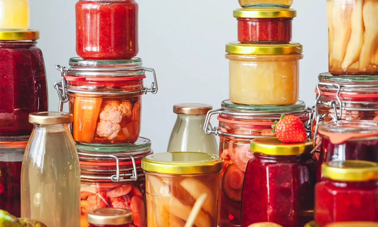 Sklenice různých velikostí s barevným obsahem od červené marmelády po světlé okurky, uzavřené zlatými a stříbrnými víčky na světlém stole