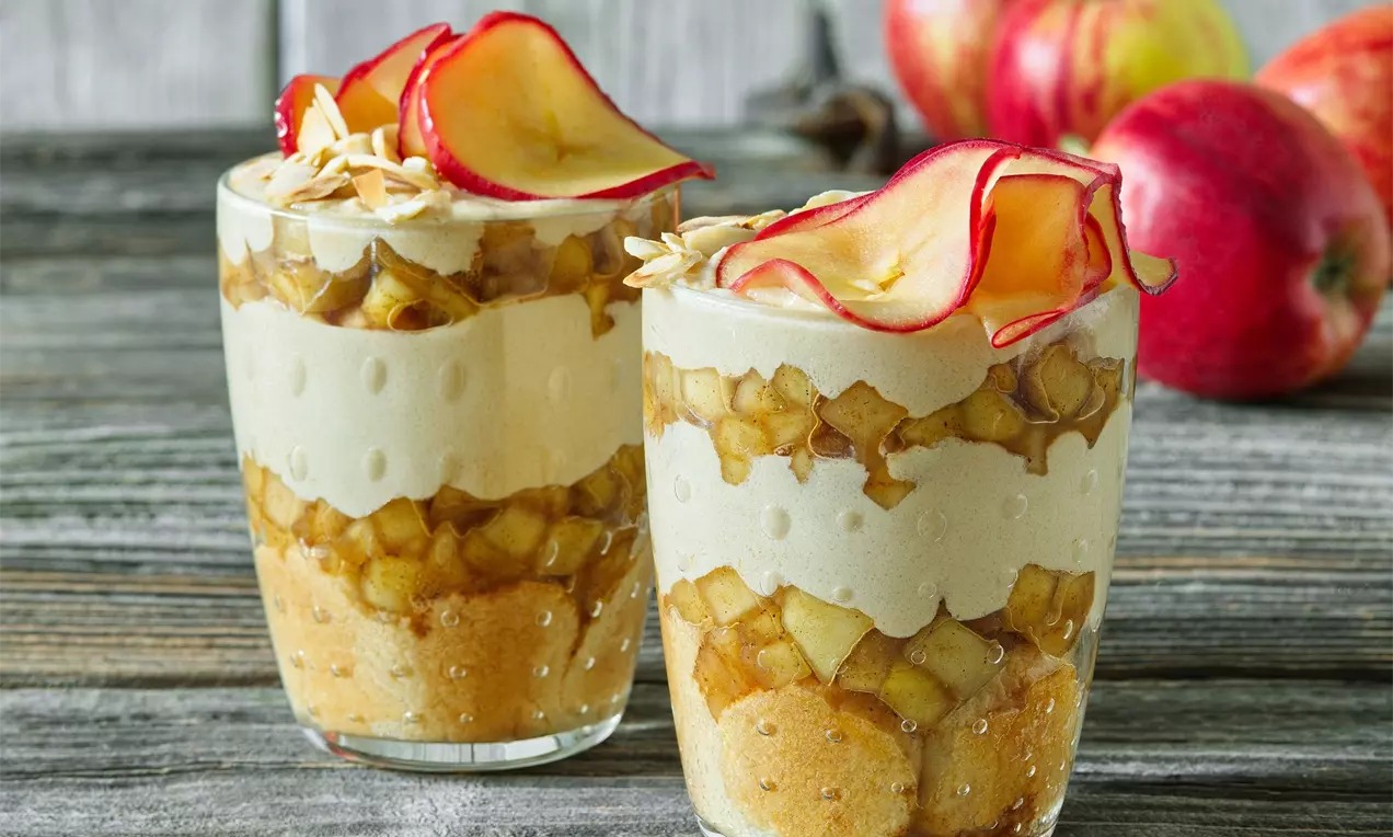 Dva vrstvené poháry z piškotů, jablek a krému, zdobené plátky sušených jablek; ve vysoké sklenici na dřevěné desce s čerstvými jablky v pozadí
