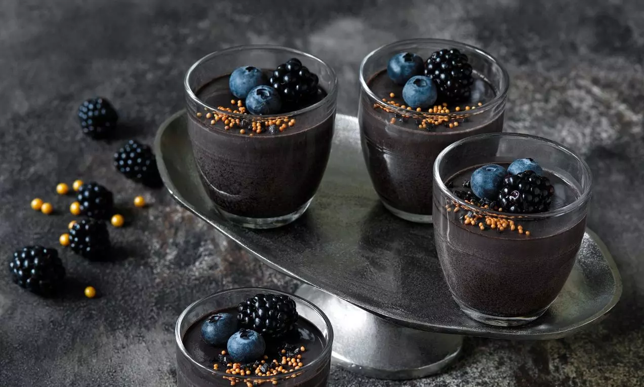 Černá panna cotta v průhledné skleničce s hladkým povrchem, zdobená čerstvými ostružinami a drobnými zlatými kuličkami na tmavém podkladu