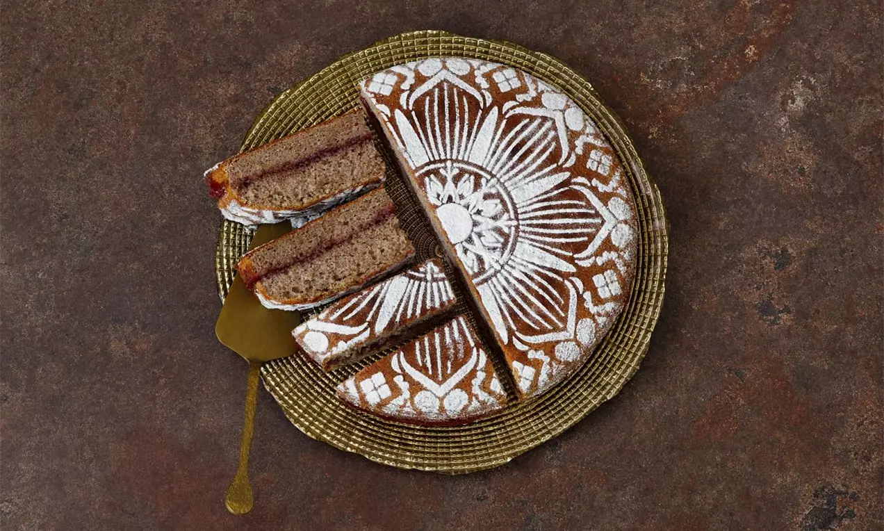 Pohankový koláč kulatého tvaru s výrazným bílým vzorem připomínajícím květinu, s několika dortovými kousky na zlatém podnosu