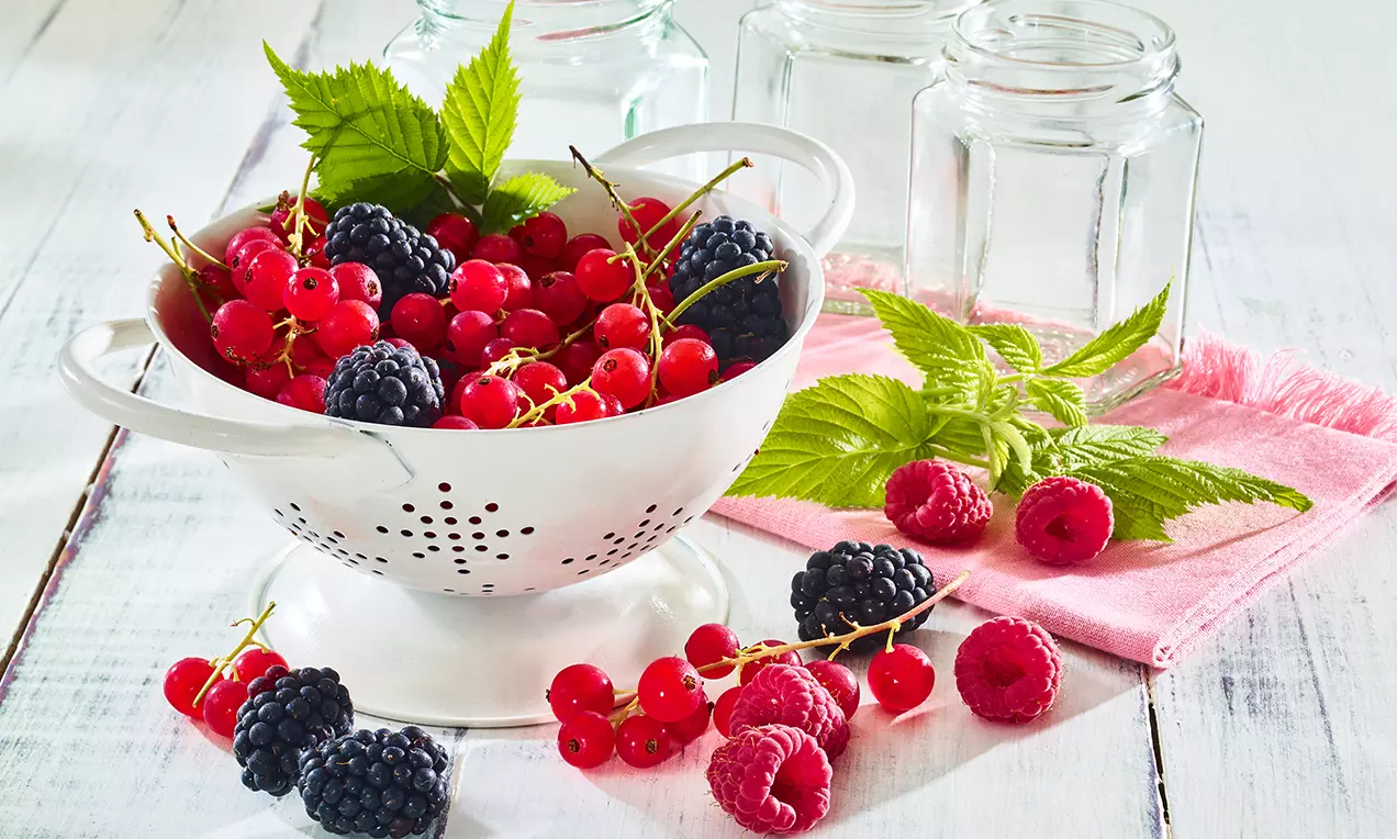 Barevné bobulovité ovoce, červený rybíz, maliny a ostružiny v bílém cedníku, doplněné zelenými lístky na světle dřevěném stole s růžovou utěrkou