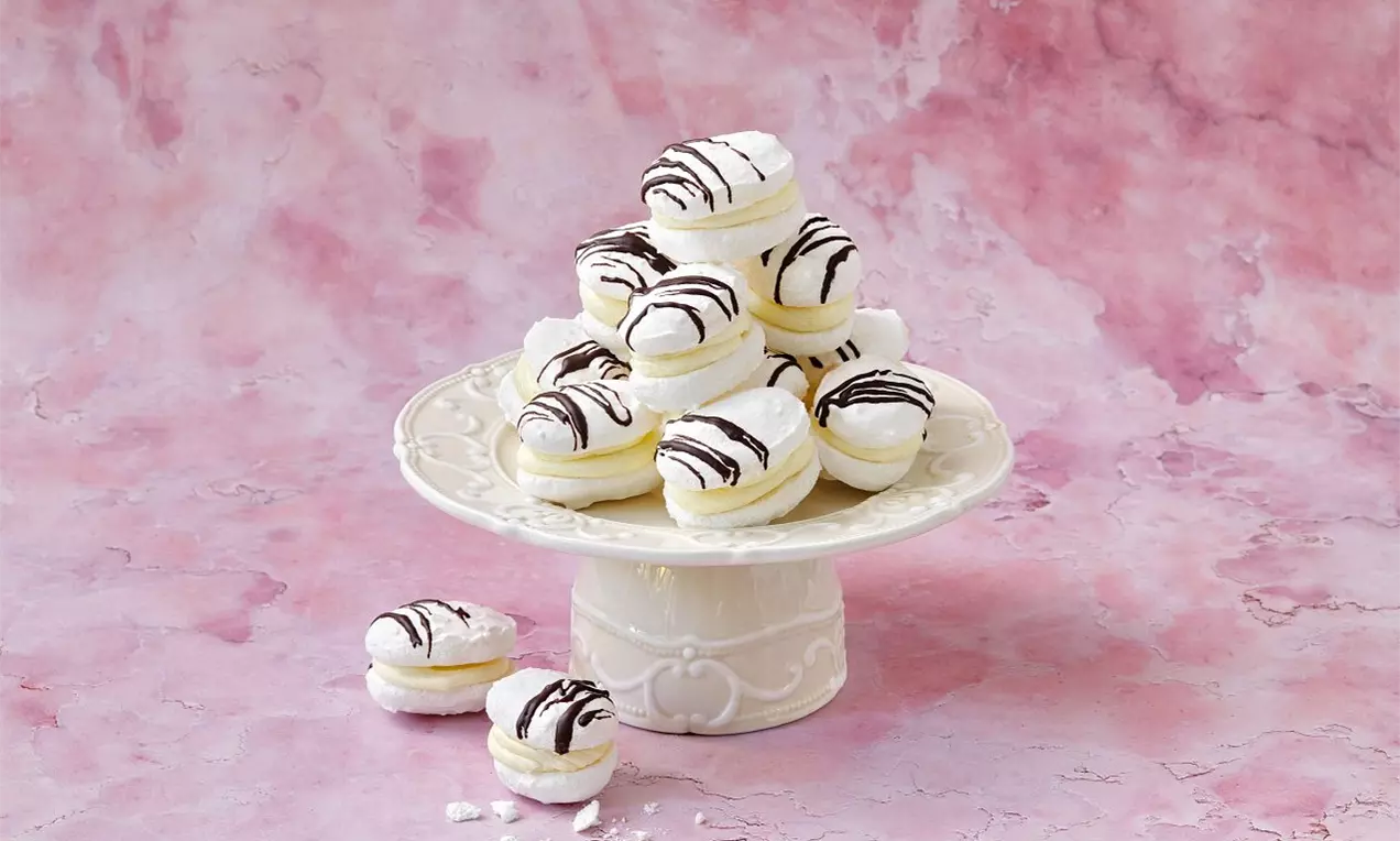 Na světlém podstavci jsou vrstvené bílé laskonky oválného tvaru s kokosovým povrchem, plněné krémem a zdobené proužky čokolády