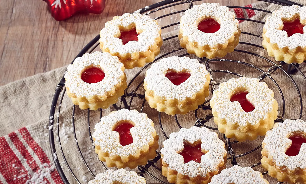 Kulaté jemně zlatavé sušenky s vykrojeným středem plněným červenou marmeládou, posypané moučkovým cukrem na černé mřížce