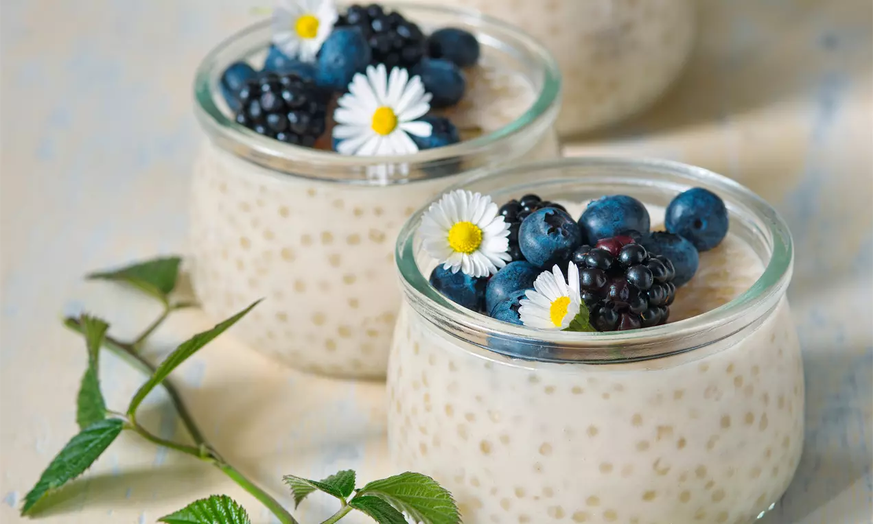 Krémový tapiokový puding v průhledných skleničkách ozdobený čerstvými borůvkami, ostružinami a bílými sedmikráskami na světlém podkladu