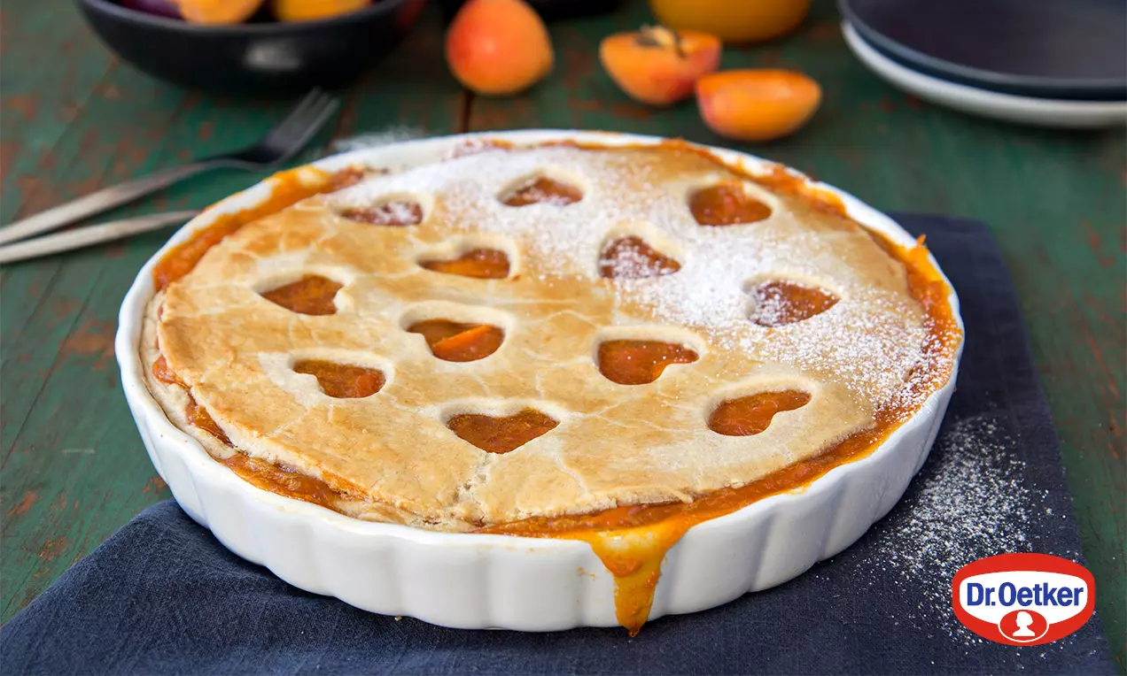 Meruňkovo-pomerančový pie se zlato-hnědou kůrkou, srdcovitými výřezy, na bílém zapékacím talíři, ozdobený moučkovým cukrem, s čerstvými meruňkami
