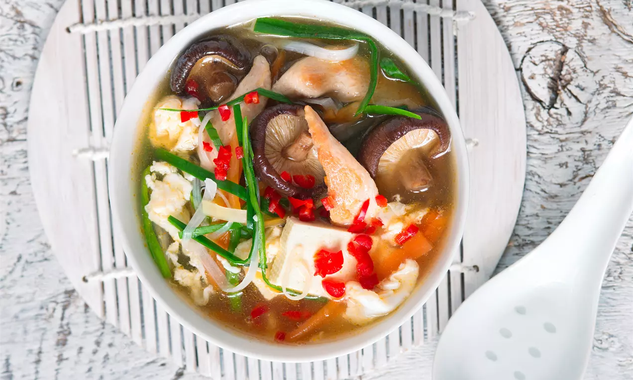 Světlý vývar s plátky masa, houbami, tofu, červenou chilli papričkou a zelenou cibulkou v bílé misce na dřevěném podkladu