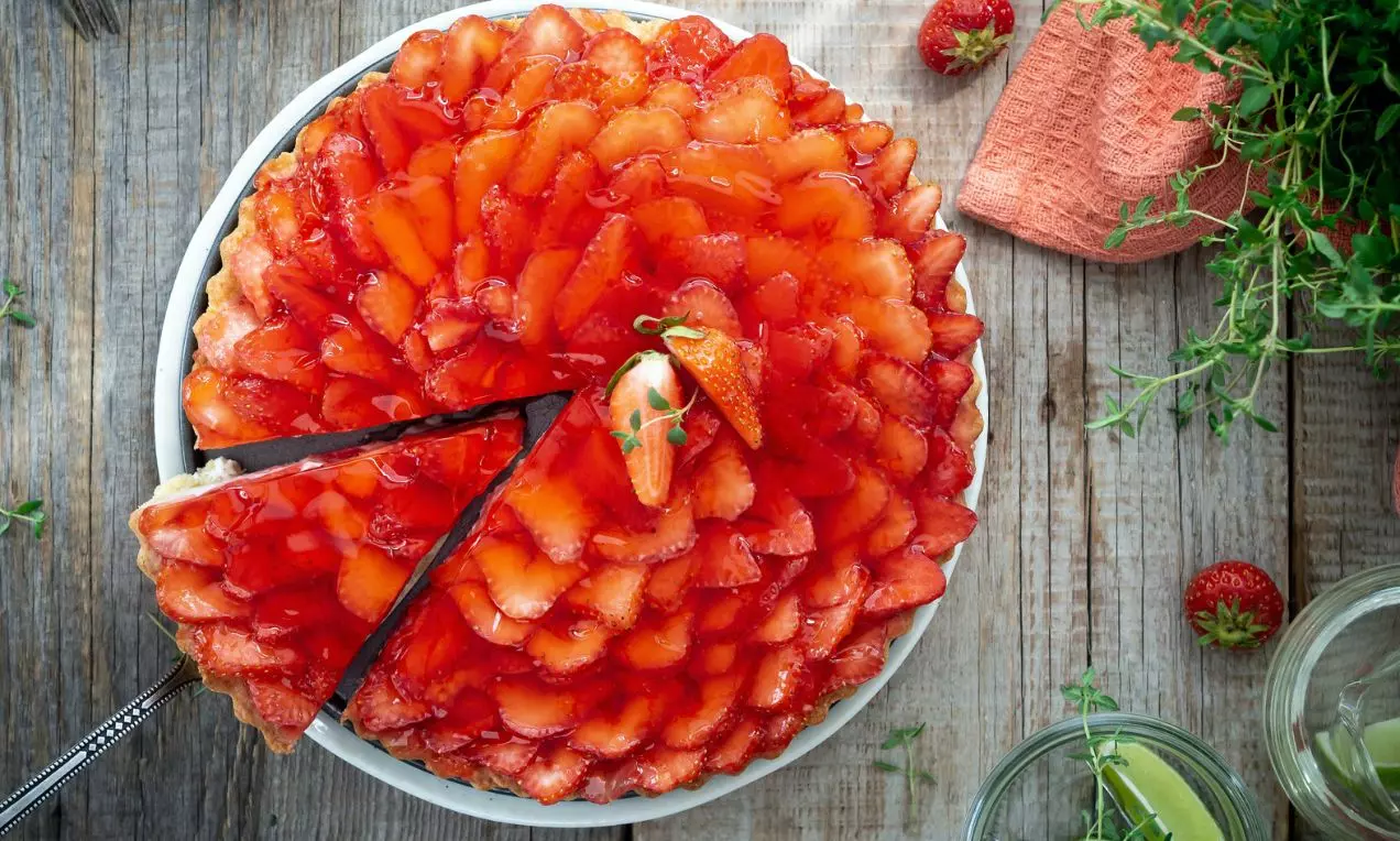 Jahodový tart s lesklými plátky jahod uspořádanými do kruhu na světlém korpusu, podávaný na bílém talíři s čerstvou bylinkou uprostřed