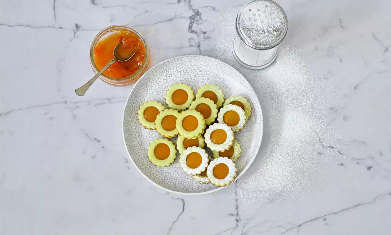 Světle zelené kytičky z lineckého těsta s hladkým povrchem, plněné zářivě oranžovou marmeládou, na bílé misce na mramorovém podkladu