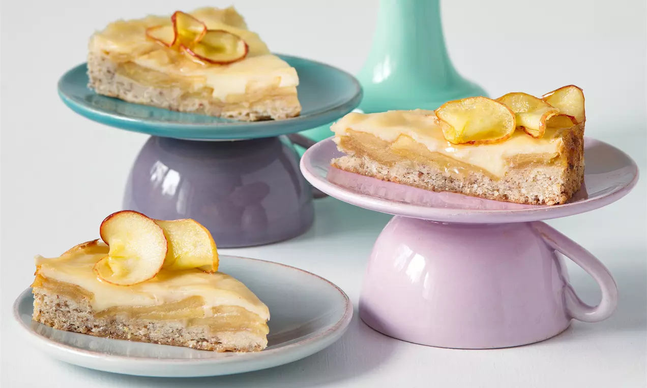 Trojúhelníkové porce světle hnědého koláče s jemnou vrstvou jablek a krémem, zdobené sušenými plátky jablek na pastelových talířích