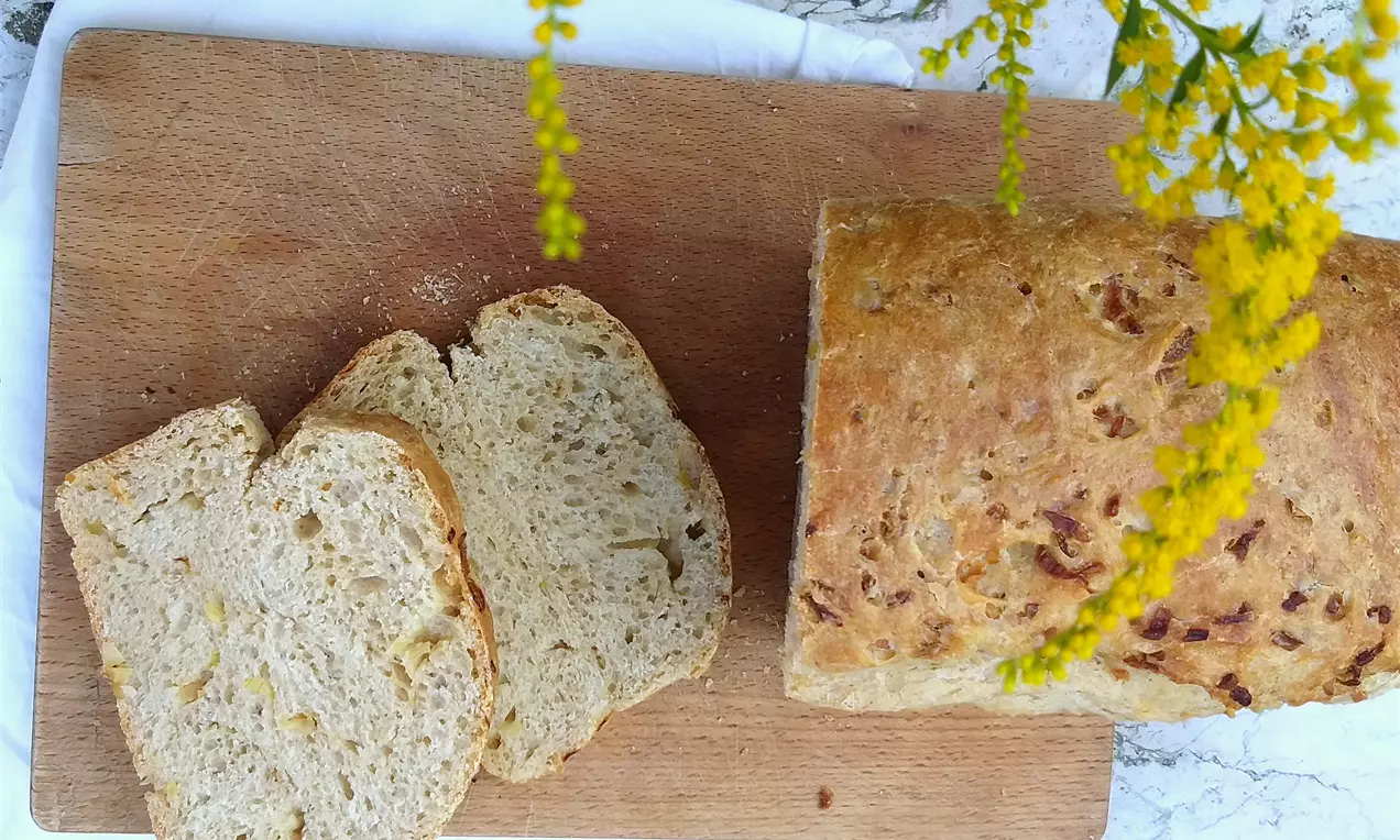 Cibulový chléb zlatohnědé barvy s křupavou kůrkou, podélně nakrájené plátky s jemnou strukturou na dřevěném prkénku s květinovou dekorací