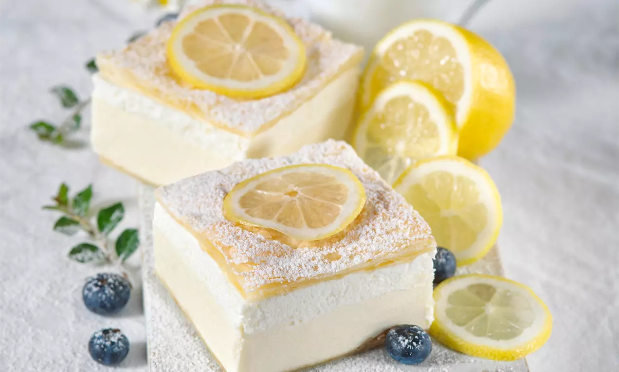 Čtverce světlého krémeše s jemnou krémovou vrstvou, posypané moučkovým cukrem, ozdobené plátkem citronu, obklopené borůvkami a citronovými plátky