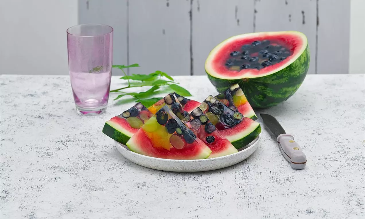 Trojúhelníkové kousky melounu plněné barevným vrstveným ovocným želé s borůvkami a plátky ovoce na bílém talíři, vedle sklenice vody