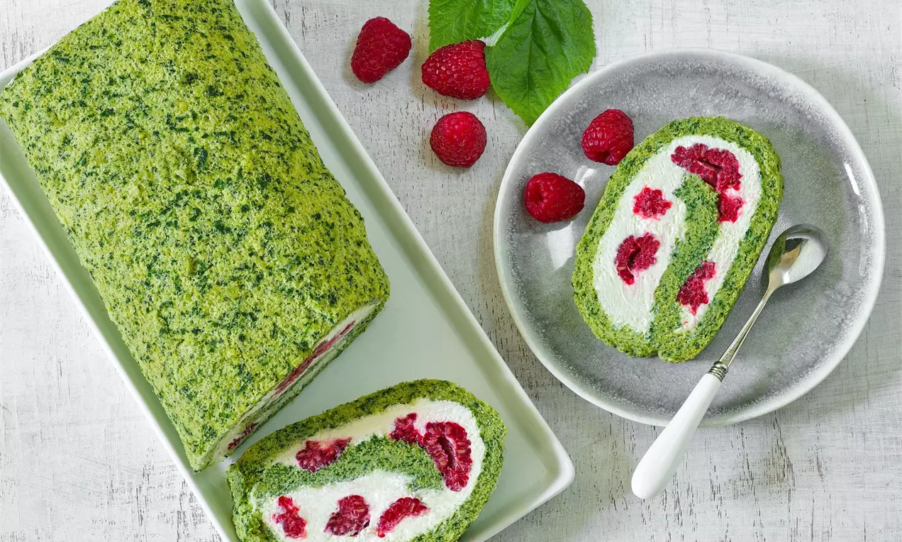 Zelená roláda plněná bílou krémovou náplní a malinami na bílém servírovacím podnose a bílém dřevěném stole, zdobená čerstvými malinami a malinovými lístky