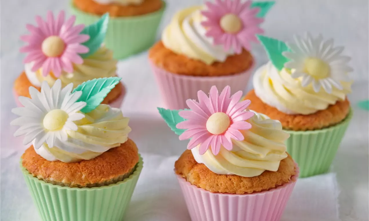 Banánové cupcakes se světlým krémem ve spirálách, zdobené barevnými cukrovými květy a zelenými lístky, uspořádané na bílém prostírání