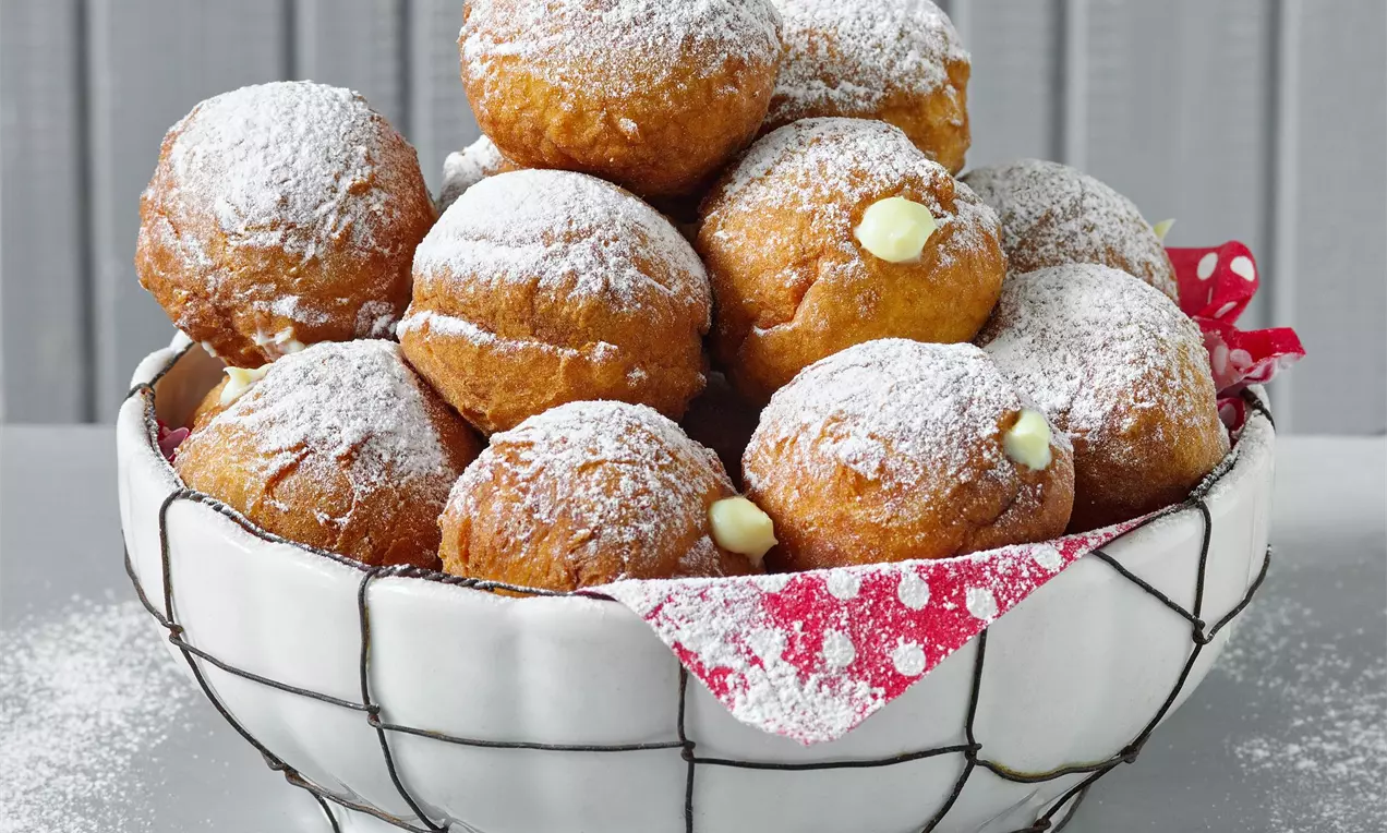 Zlatavé kulaté tvarohové koblížky lehce poprášené moučkovým cukrem, naskládané v bílé košíkové misce s látkou s červeným vzorem